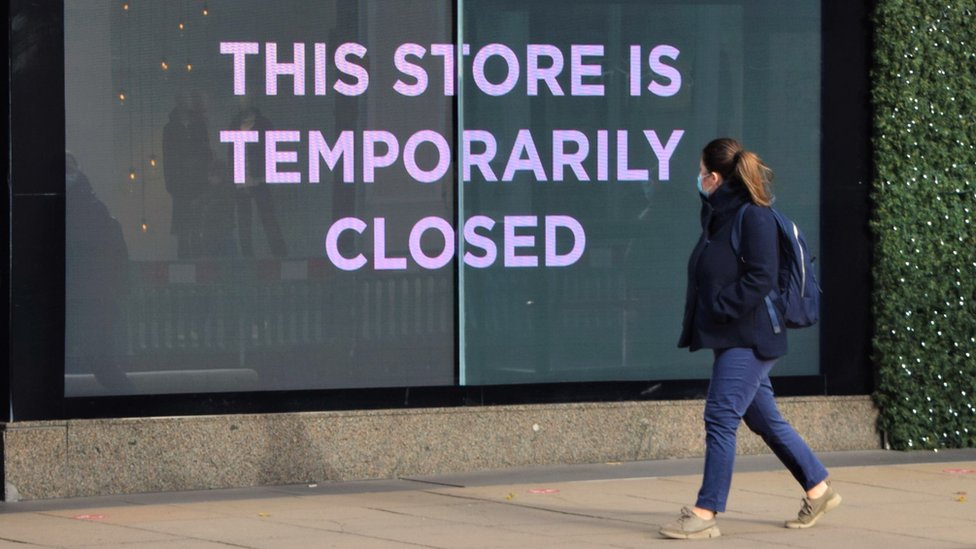 Женщина в маске проходит мимо вывески магазина с надписью «Этот магазин закрыт»
