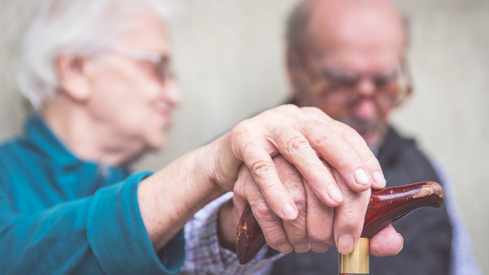 Una de las cosas tristes del Alzheimer es que los pacientes dejan de reconocer a sus seres queridos. Dos ancianos sentados, él con bastón y ella cogiéndole una mano.