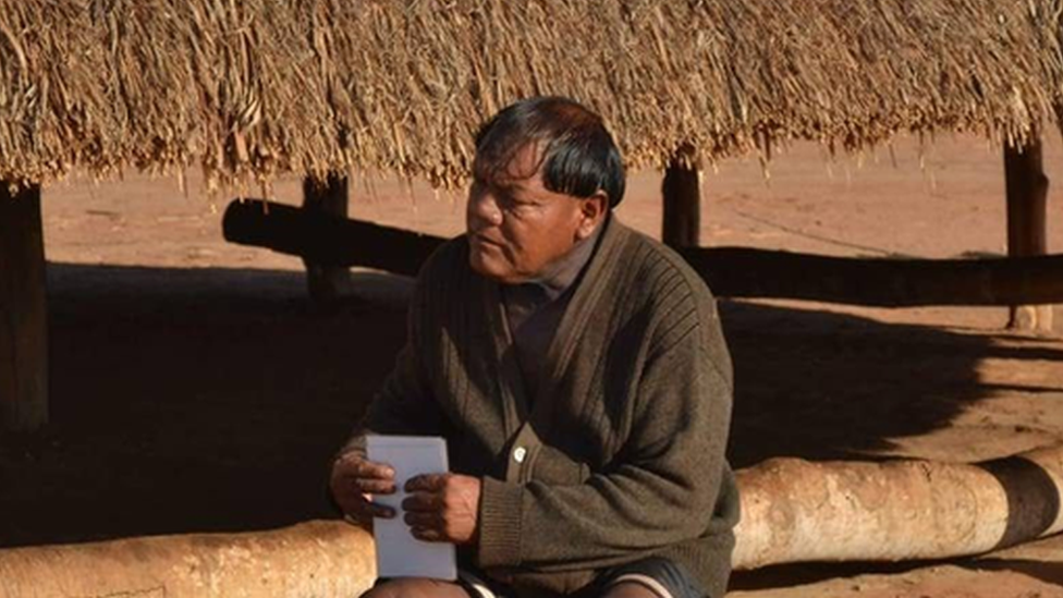 Aritana Yawalapiti, líder do Alto Xingu, sentado em frente a uma construção tradicional