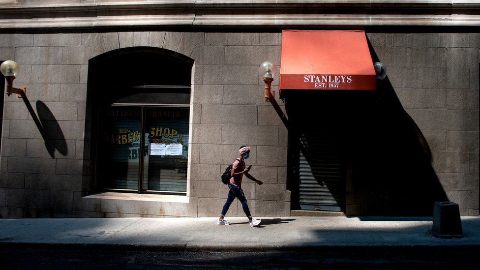 A woman, wearing a face mask, walking down a New York street and a barber's with the shutters down