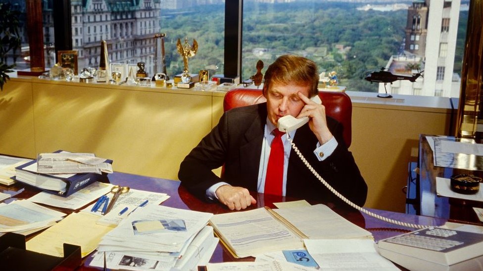 Donald Trump talking on the phone behind a desk at the top of Trump Tower 