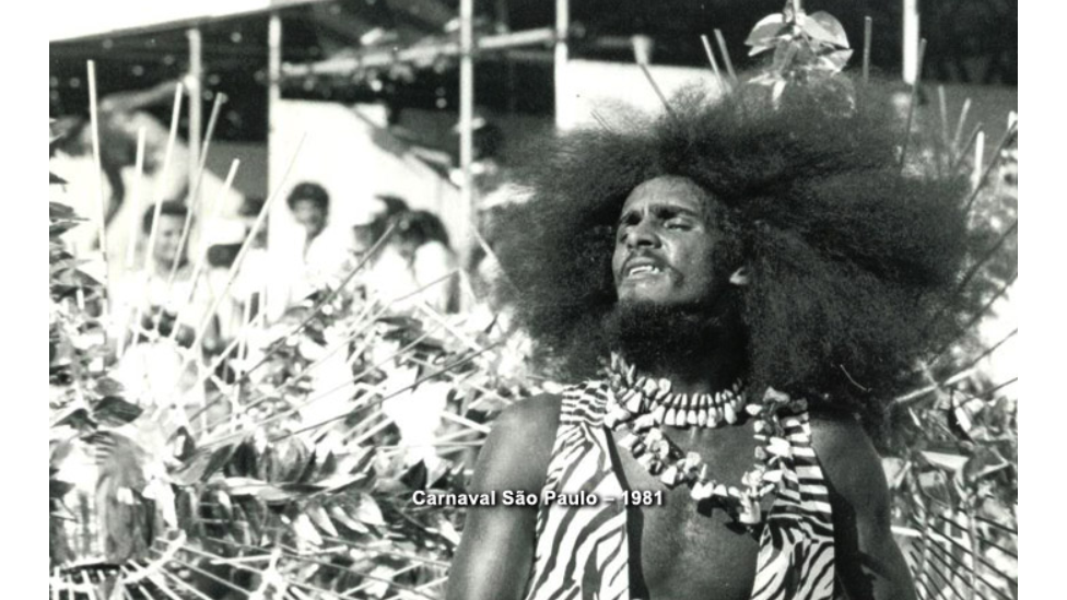 Nelson Triunfo com um enorme cabelo black power, de olhos fechados e com roupas que remetem à África, durante um desfile de escola de samba