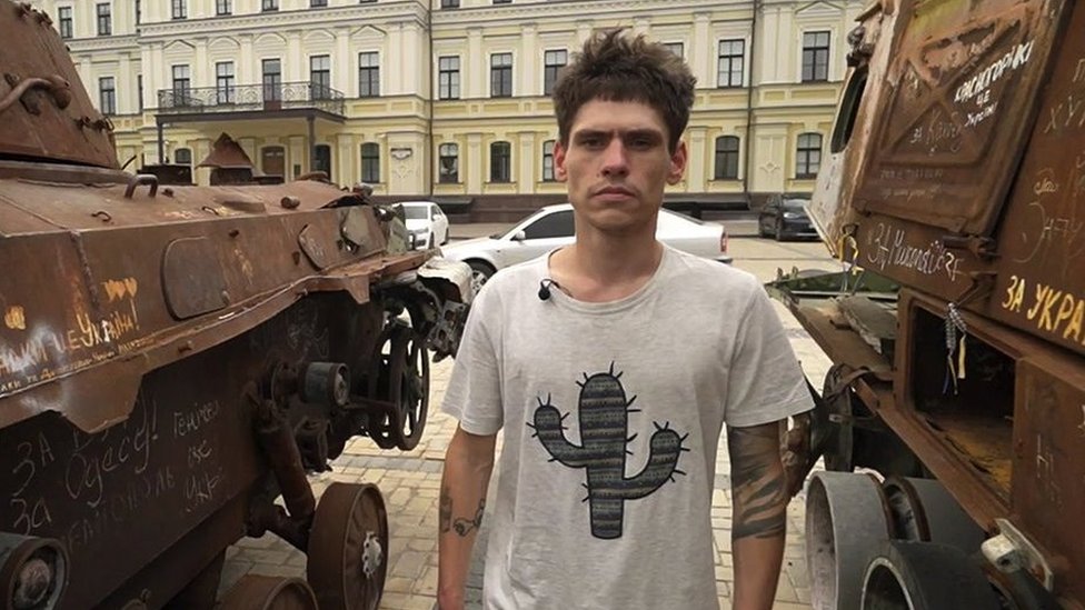 Oleksii standing between two military vehicles in Kyiv after his rescue. He has short brown hair and is wearing a white T-shirt with a picture of a cactus on it.