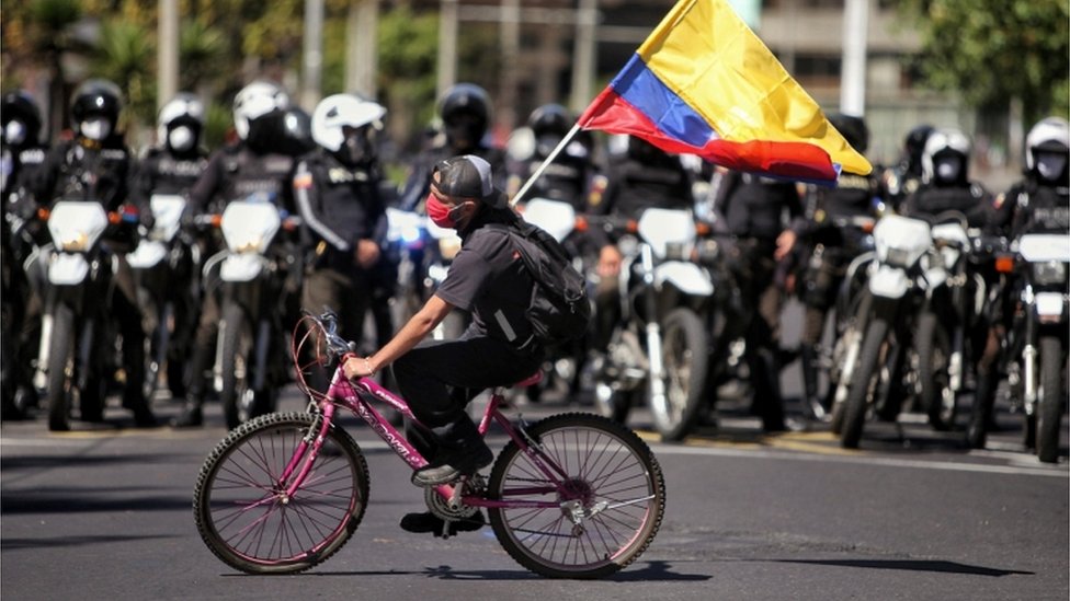 Протестующий на велосипеде машет флагом перед вереницей полицейских в Кито