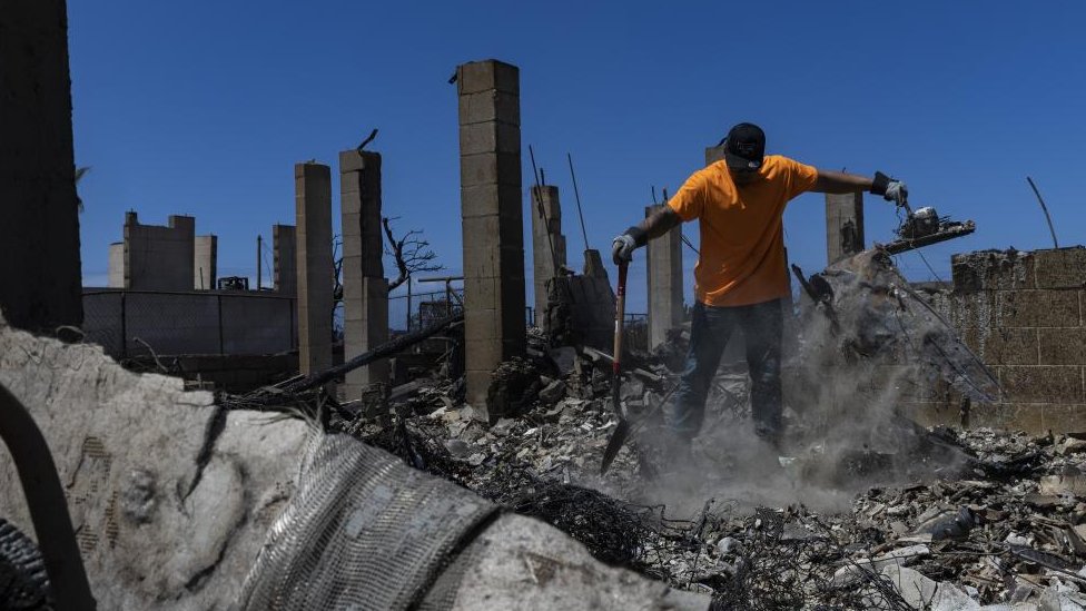 A man with a shovel stands on a large pile of debris in the ruins of a house