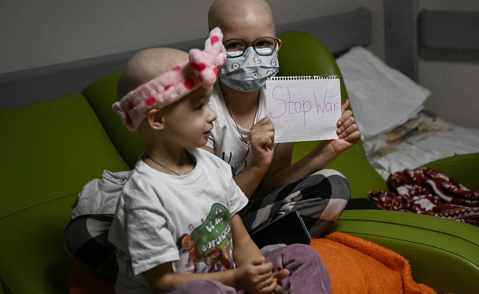 Dos niñas que reciben tratamiento oncológico, una con una moña en la cabeza, con un cartel que dice "paren la guerra"