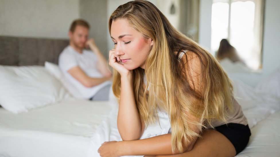 Mujer sentada en el borde de una cama tras una aparente disputa con su compañero