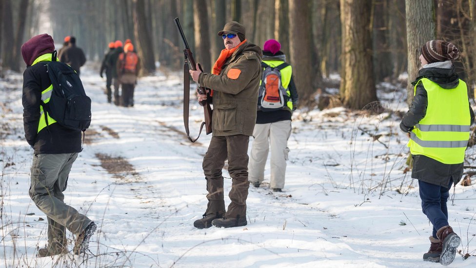 Lovac sa puškom gleda okolo dok aktivsti za prava životinja obučeni u vidljive žute prsluke šetaju okružujući ga sa svih strana