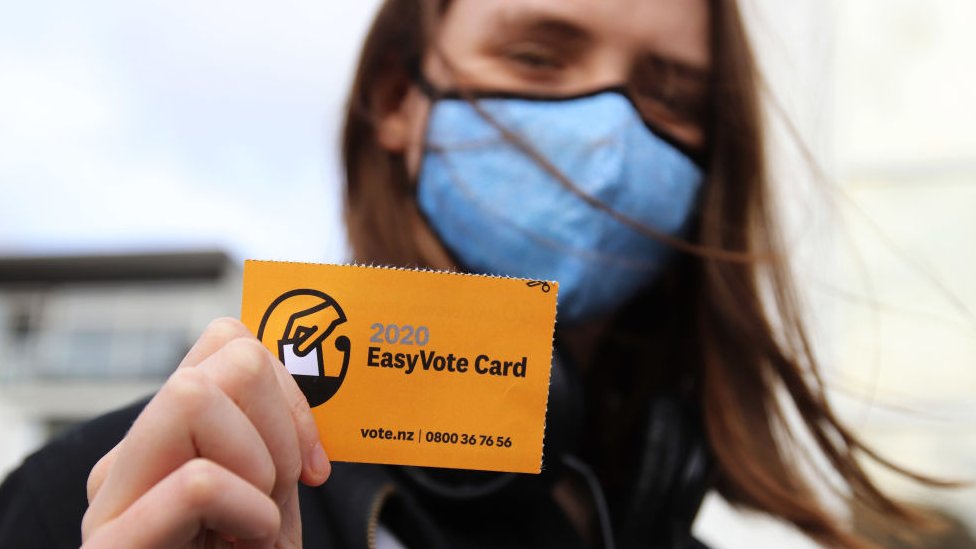 A young woman wearing a face mask holds up her Easy Vote card on the way to vote on the first day of advance voting