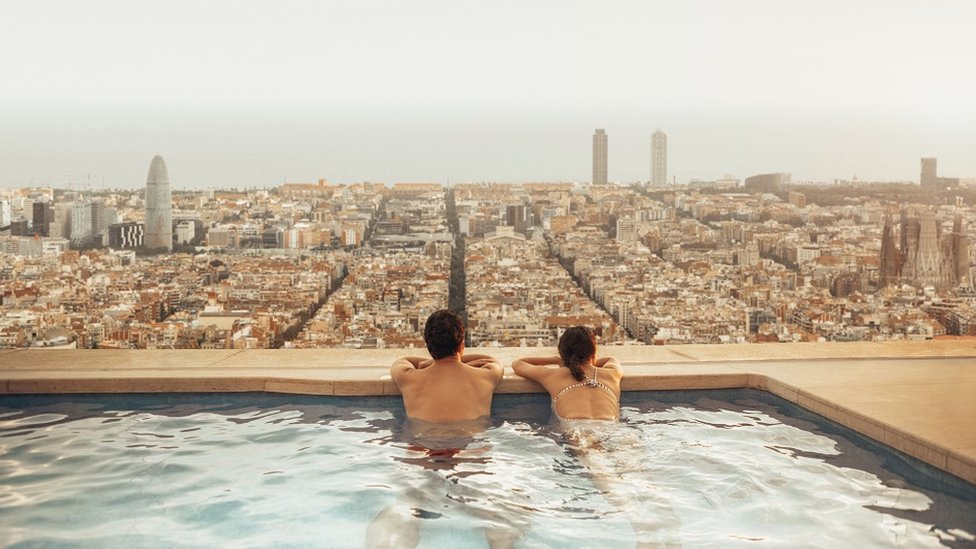 A couple on a rooftop pool in Barcelona. It's a cloudy day, and they are looking down over the city from inside an infinity pool.