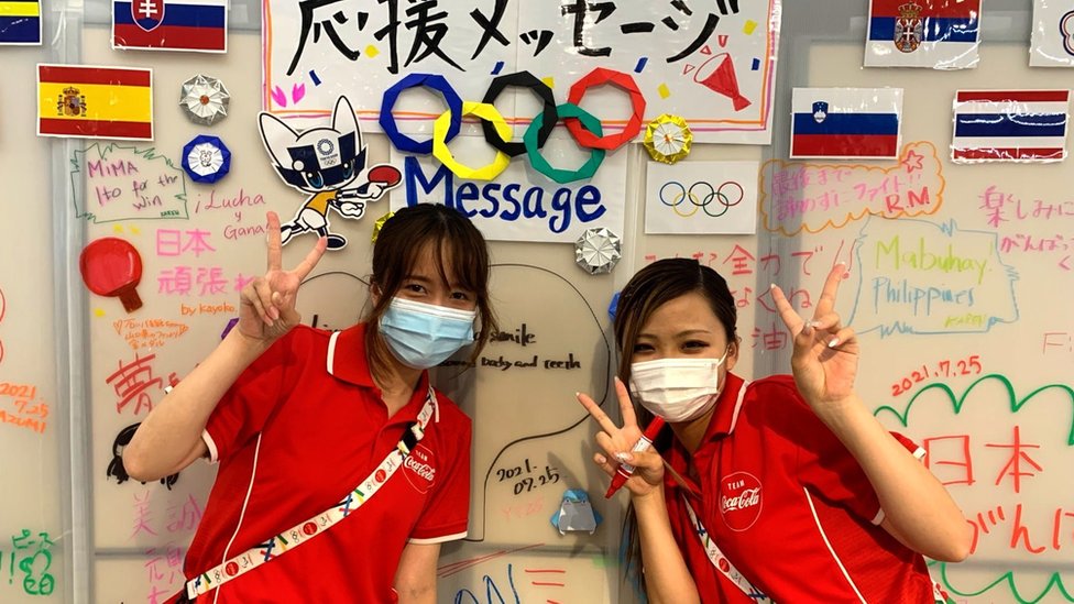 Two volunteers pose behind a wall with handwritten messages to cheer on Olympic athletes