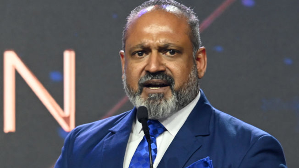 Jack Devnarain wearing a blue suit and tie speaks at a microphone.
