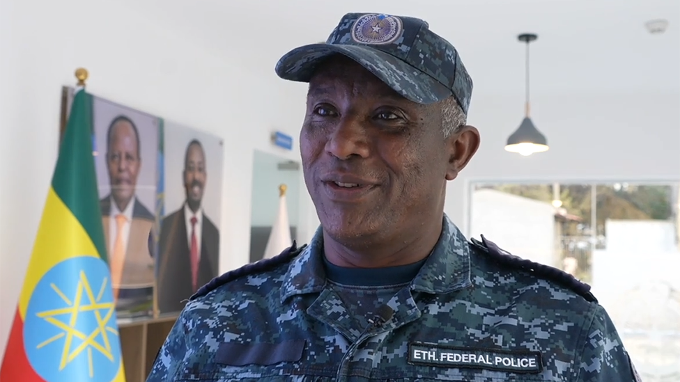 A head and shoulders image of a man in a blue camouflage uniform and peaked cap. A badge says "Eth Federal Police" on his chest and an Ethiopian flag is in the background.