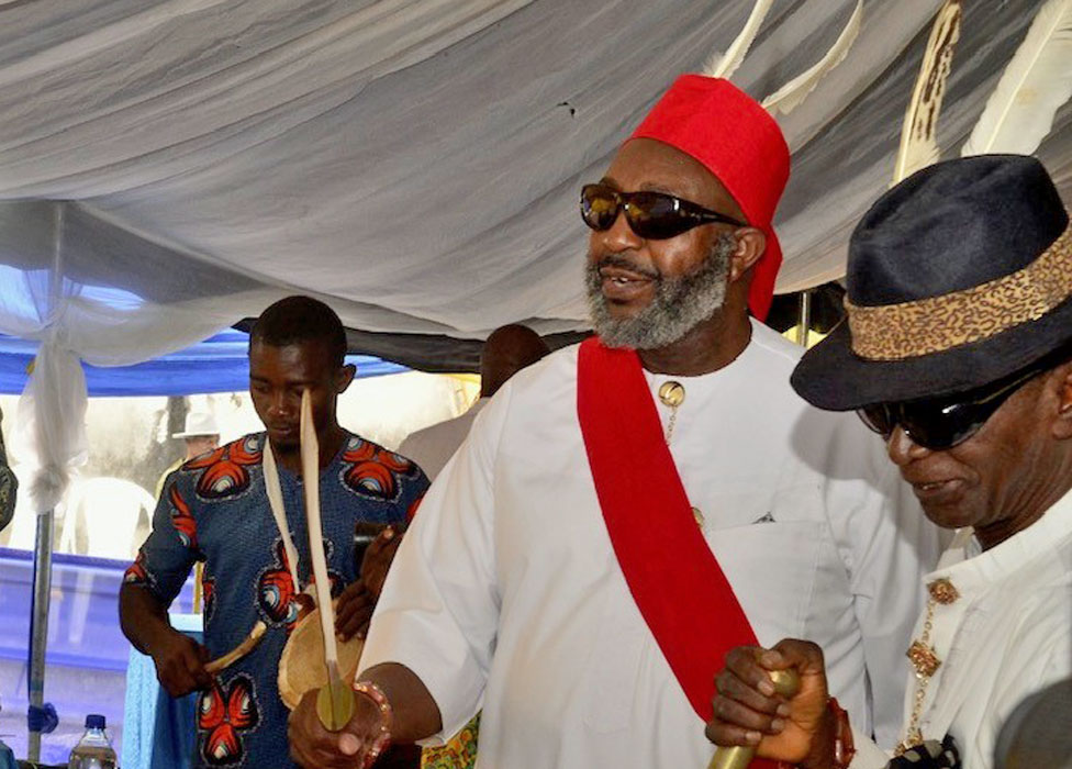 Ken Okoroafor in sunglasses and wearing a white robe, red sash and red hat and holding a gold sword