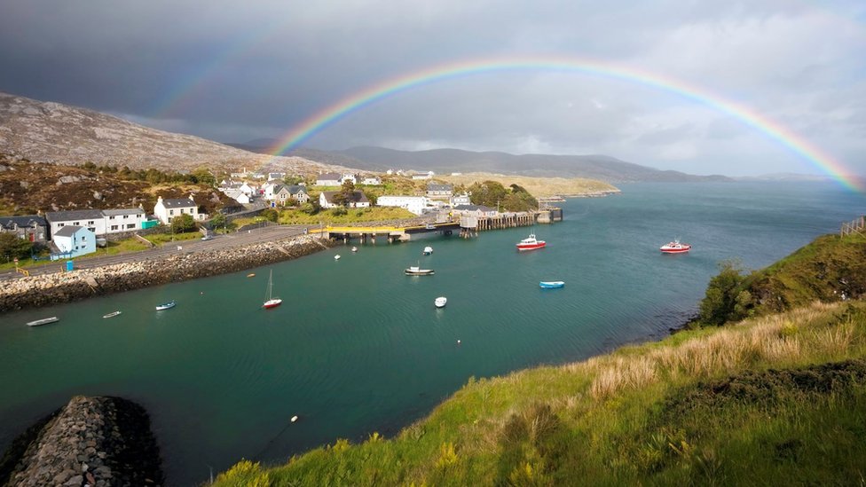 A rainbow arches over a sea loch and Hebridean community. There are several boats in the water.