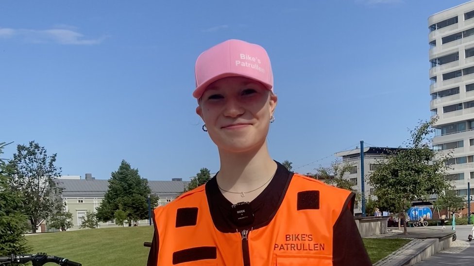 Olivia Hamalainen wears a pink baseball cap and orange high vis vest standing outside a block of flats.