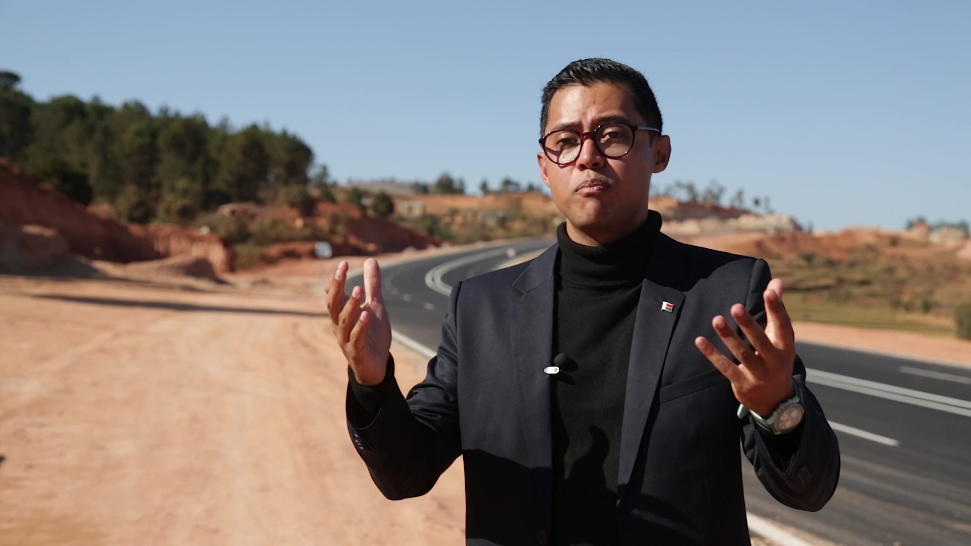 A man in a dark jacket and black polo neck top gesticulates as he stands by the side of a new road.