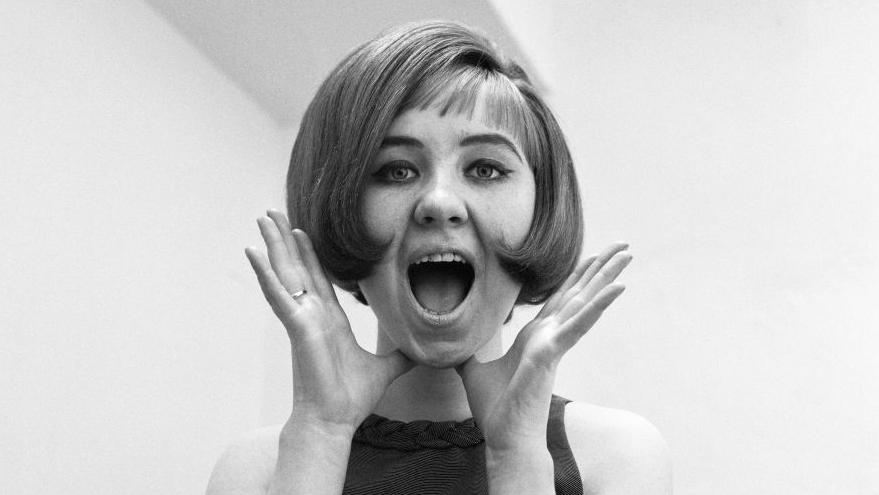 Lulu pictured in 1964. She has her hands raised to her face and is singing.