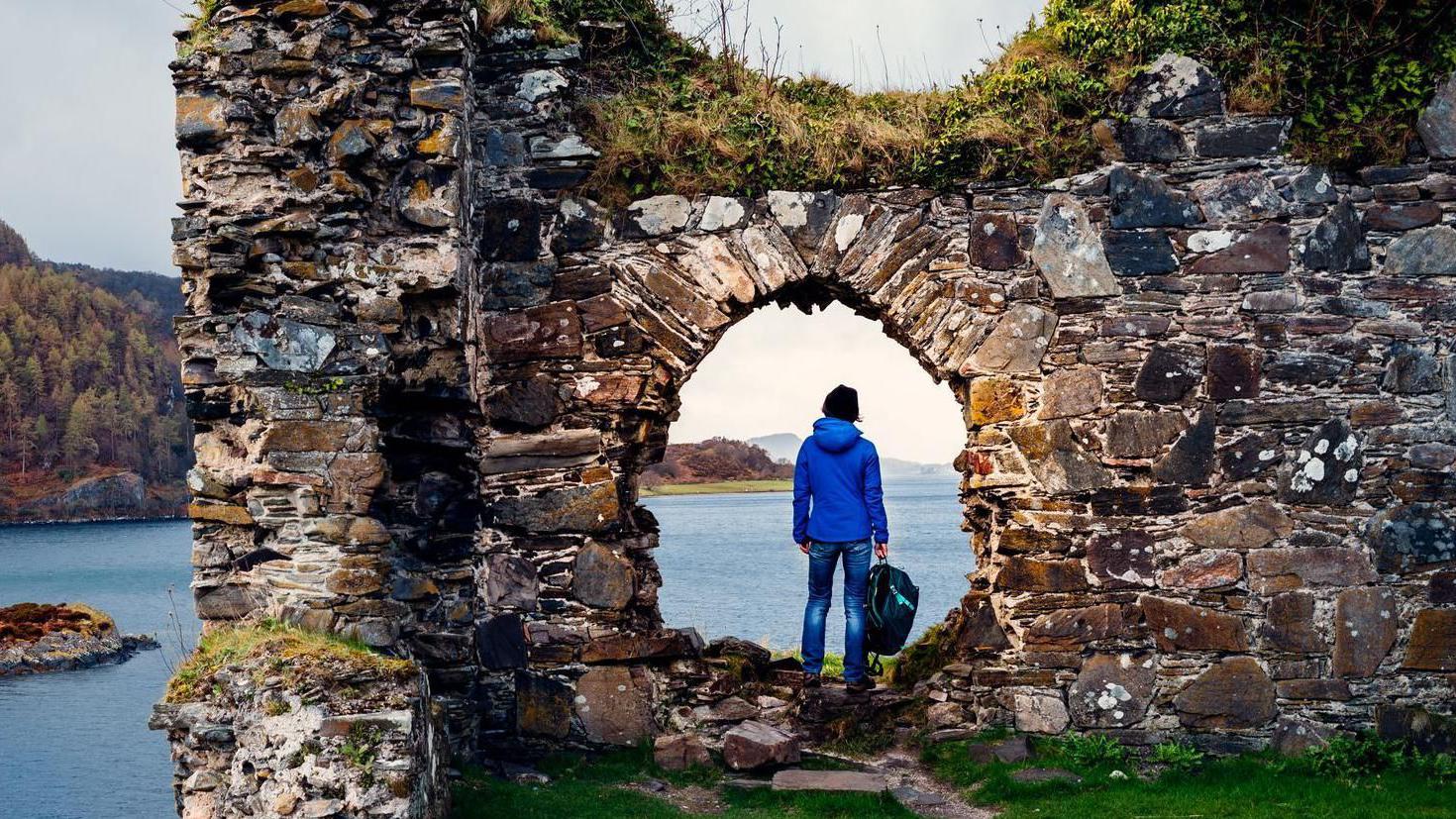 A person stands in an arch in a wall of Strome Castle. The ruins are capped by grasses and moss and overlook a loch.