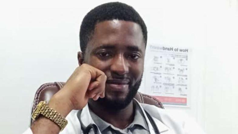 Augustine Ogbo smiles at the camera with his face rested on his hand. He wears a watch, a white doctor's jacket, and a stethoscope around his neck.