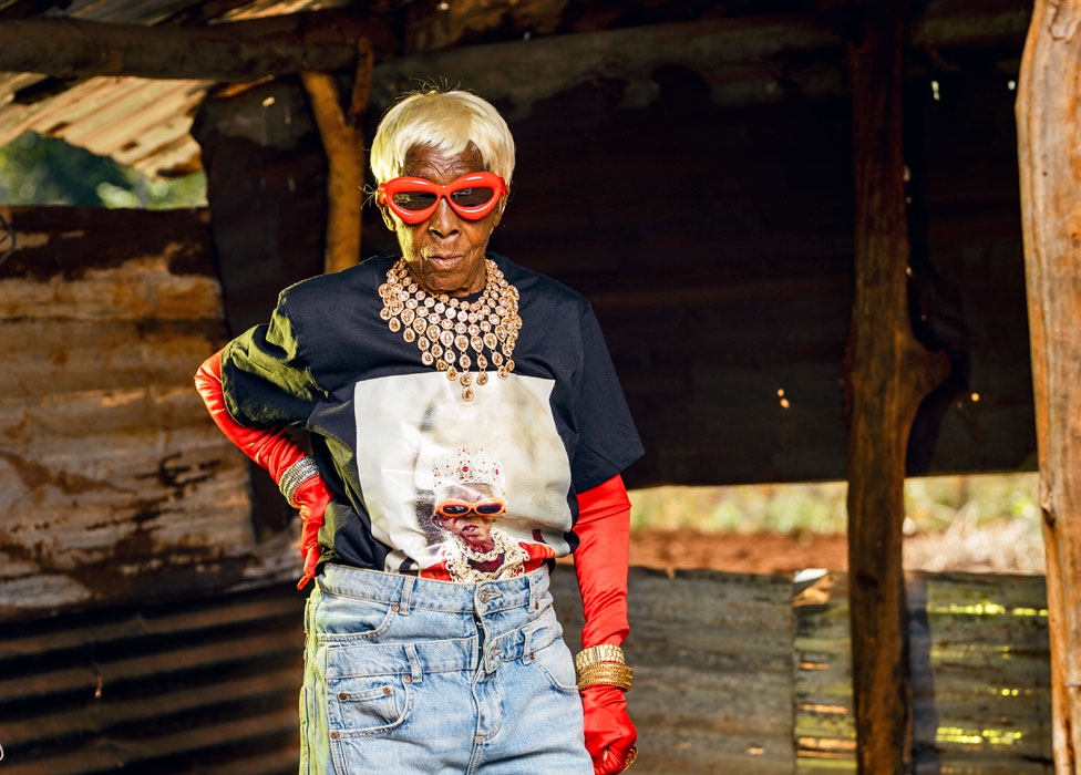 Margret Chola in jeans, a blond wig and red gloves. She also wearing a T-shirt with her image on the front, big jewelled necklace and red sunglasses