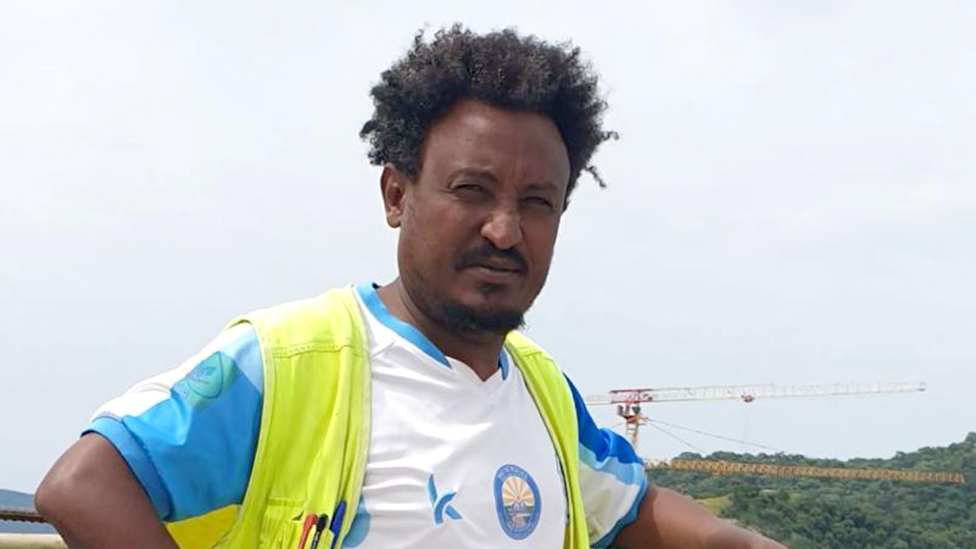 Moges Yeshiwas squinting into the camera. He is wearing a sports T-shirt and a hi-viz waistcoat. A crane can be seen in the background.