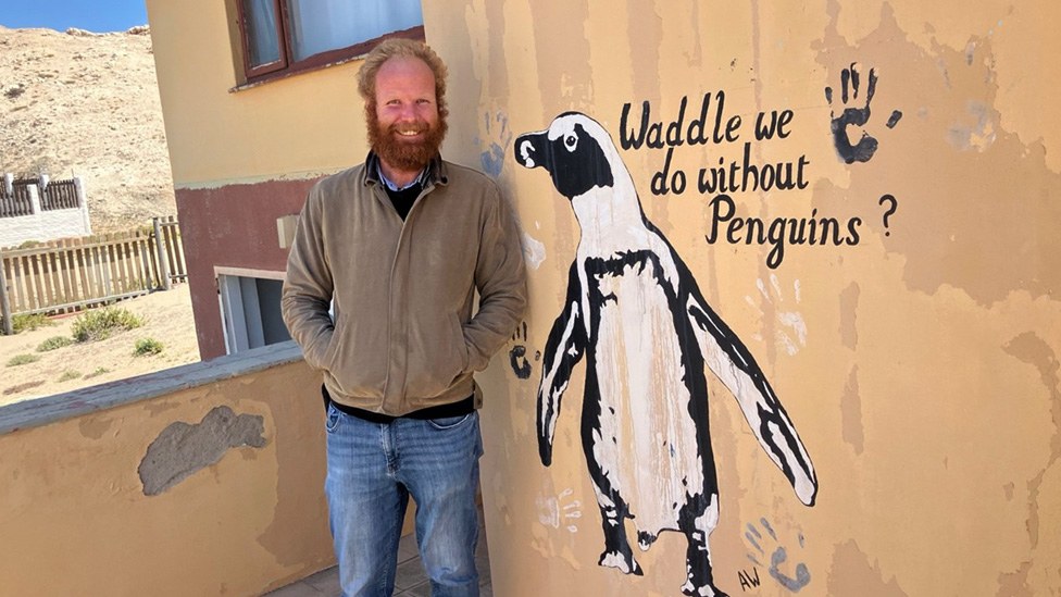 Neil Shaw, operations manager for Namcob stands on a veranda outside his office, next to a wall painting, depicting a penguin and the inscription ‘Waddle we do without Penguins?