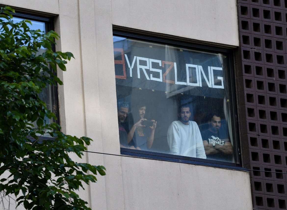 Four asylum seekers and refugees held inside a Melbourne hotel stand at a window with the sign '9 Yrz 2 Long'