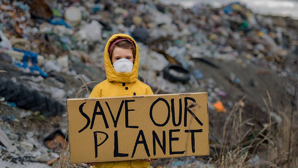 Niña frente a un vertedero de basura con un cartel que dice "salvemos a nuestro planeta"