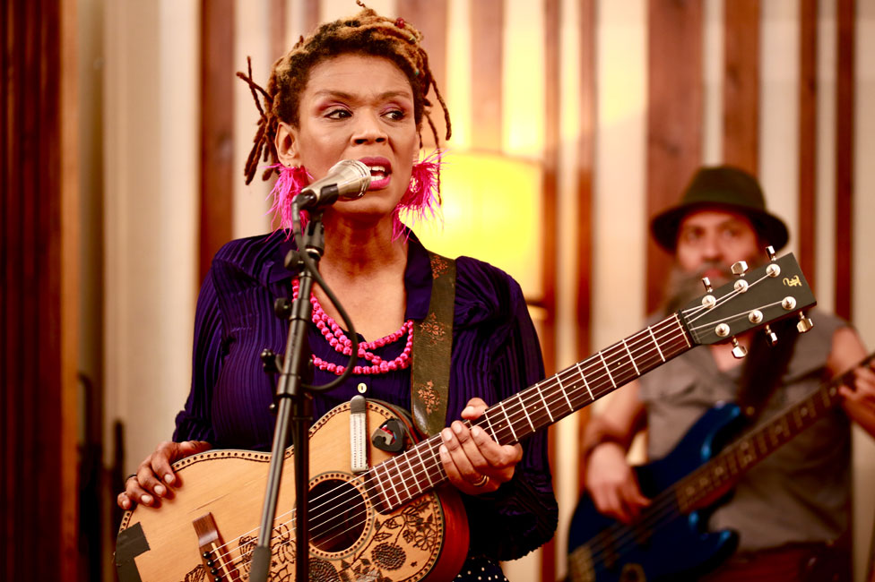 Carmen Souza singing, holding a guiltar, another guitarist can be seen behind her