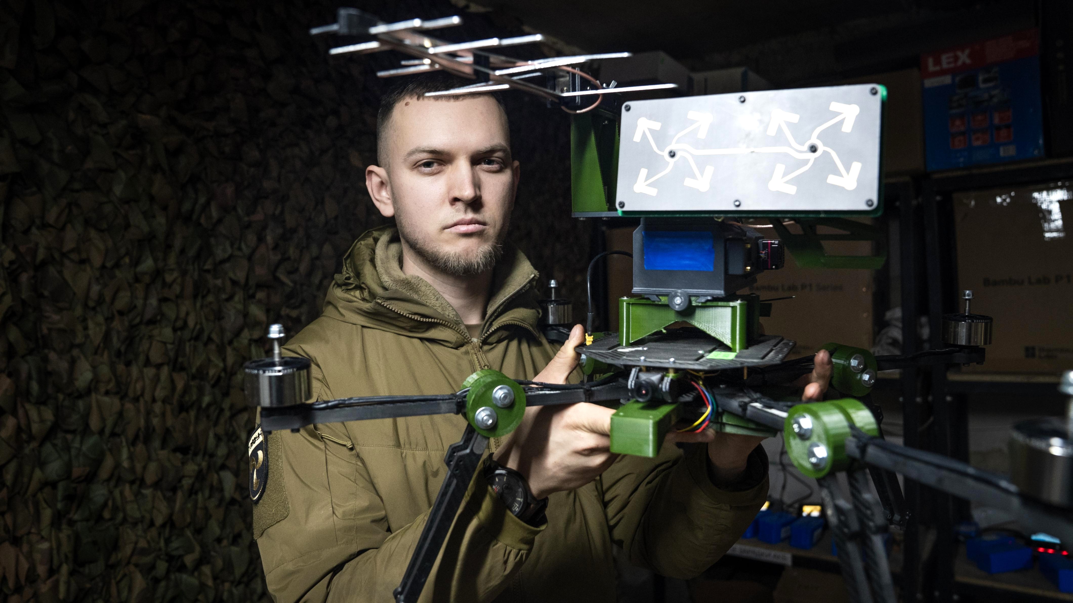 Roman, a Ukrainian soldier in a drone repair unit, holds up an unmanned aerial vehicle and faces the camera. He's in his green army uniform.