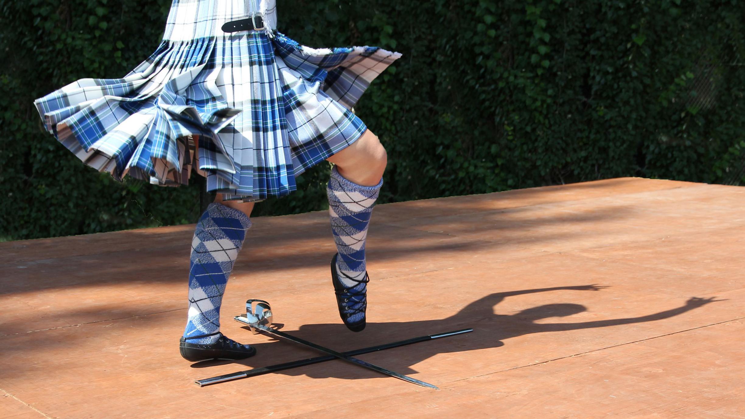 A Highland dancer wearing kilt and knee length socks of blue and white tartan performs a sword dance.