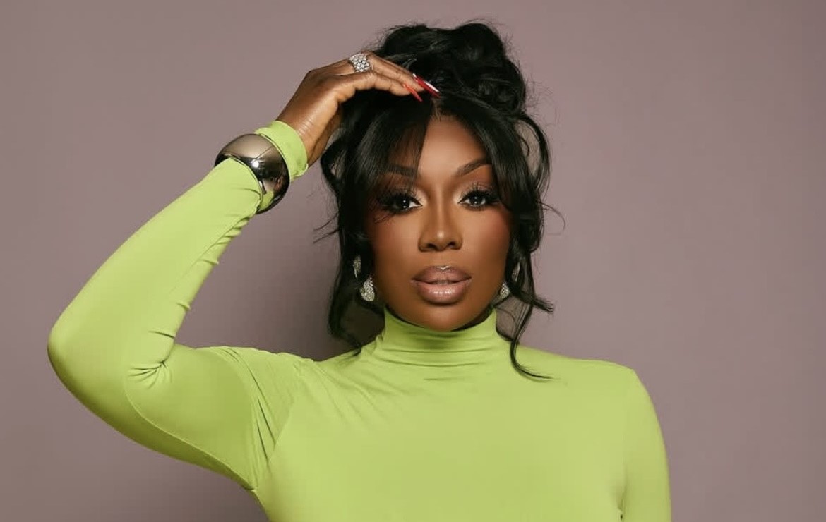 A woman wearing a green top poses with her left hand touching her hair.