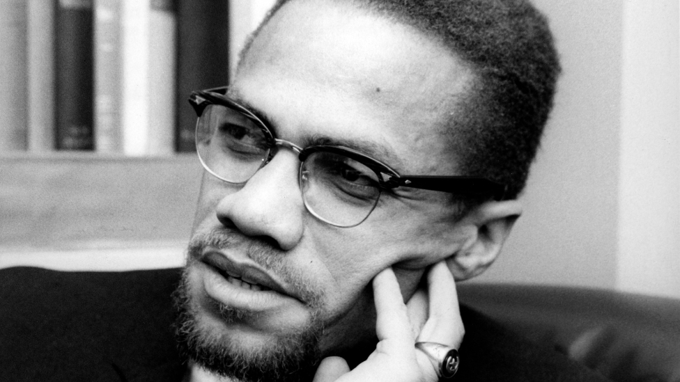 Malcolm X, pictured at Broadcasting House. He is wearing a black suit, black rimmed glasses and a ring on his finger.