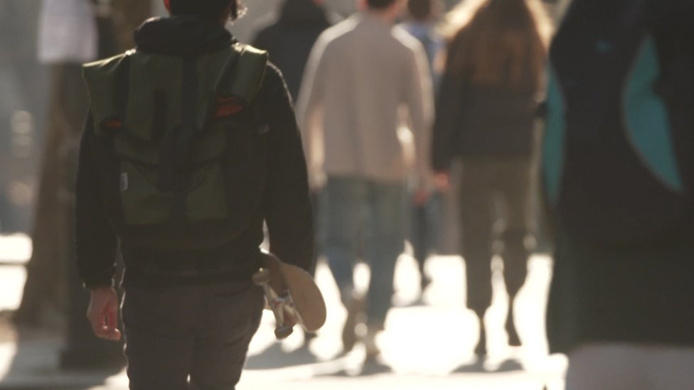 An anonymous student carrying a skateboard walks behind groups of other anonymous students.